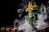 A tiki cocktail in a detailed green cup, garnished with a sliced citrus fruit and a gooseberry, surrounded by swirling smoke, sits on a rustic wooden surface in a dimly lit setting