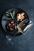 Top view of a sophisticated arrangement with Greek BBQ olives in a small black bowl, a portion of crumbled feta cheese flavoured with herbs, a glass of dry white wine and fresh sage leaves on a dark, textured surface. Ideal for a Mediterranean gourmet starter.