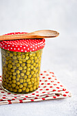 A jar of preserved homemade organic peas with a red dotted lid is topped with a wooden spoon. The jar stands on a dotted cloth and offers a lively country-style kitchen scene.