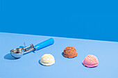 Three scoops of ice cream in chocolate, strawberry and vanilla flavours on a bright light blue and blue two-tone background, accompanied by a metal scoop with a blue handle.
