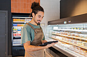 A café employee in an apron works with a tablet while standing in front of a display of pastries The scene shows a modern, efficient coffee shop