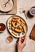 Top view of a cropped, unrecognisable hand holding a ceramic plate of sugared churros, with bowls of cinnamon and rich chocolate sauce beside it, taken in a domestic kitchen with a natural linen background.