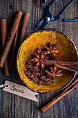 Top view of a rustic yellow bowl filled with star anise and cinnamon sticks, accompanied by an old pair of scissors and a label with the words Spices. Ideal for illustrating cosy, aromatic culinary themes.
