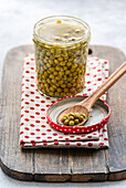 A jar of green, homemade organic peas is presented on a rustic wooden board with a wooden spoon of organic peas on top. A red polka-dotted cloth adds a cosy touch.