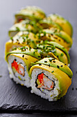 Close-up of a sushi roll, topped with fresh avocado slices, sprinkled with sesame seeds and garnished with chopped chives on a black slate plate.