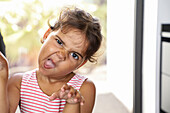A playful child in a striped top makes a silly face and gesticulates dramatically while indoors. The natural light emphasises the lively expression and captures the youthful joy.