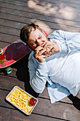 A person enjoys a barbecue meal outdoors, lying next to a skateboard. It is accompanied by chips with ketchup and a refreshing drink, creating a relaxed, laid-back atmosphere.