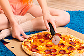 A person sits cross-legged on a blue carpet, wearing cosy clothes and slicing a delicious pepperoni pizza with a pizza cutter - perfect for relaxing or for casual dinner themes