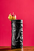 Ein auffälliger Tiki-Cocktail, serviert in einem schwarzen Becher mit einer verzierten Garnitur. Der leuchtend rote Hintergrund unterstreicht die exotische Präsentation und macht ihn perfekt für Themenpartys oder Bar-Aktionen