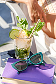 A refreshing mint and lime cocktail sits alongside sunglasses and a purple book, capturing the essence of a relaxing sunny day outdoors.