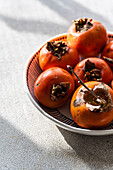 A bowl of freshly picked, ripe persimmons on a light-coloured textile surface, bathed in soft sunlight that highlights their bright orange hue and soft texture.