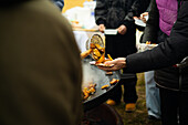 Cut-off, unrecognisable hands serving fried potatoes in a paper bowl at a lively street party where a crowd is gathered to eat.