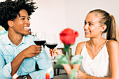 Ein fröhliches Paar stößt bei einem romantischen Abendessen mit Weingläsern an und zeigt so Glück und Zweisamkeit. Eine rote Rose unterstreicht die intime und fröhliche Atmosphäre.