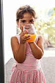 A young girl in a striped dress enjoys a refreshing glass of orange juice outdoors. The bright sunlight and the greenery in the background give the scene a summery feel.