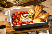A savoury dish with baked Camembert cheese, garnished with herbs, surrounded by roasted cherry tomatoes, garlic and crusty bread, served in a ceramic bowl.