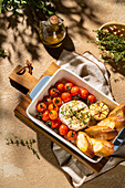 A rustic setting with baked Camembert cheese in a bowl with cherry tomatoes, garlic, herbs and crispy slices of bread on a wooden board. Ideal for gourmet cuisine themes.