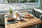 A croissant, French toast with berries, a teapot and two cups of coffee sit at a sun-drenched café table. A cosy, inviting ambience