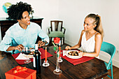 A couple enjoy a romantic dinner at home, with wine, red candles, a rose and a wrapped gift. The cosy atmosphere is perfect for a special occasion.