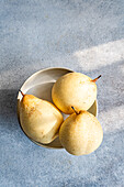Three ripe white Chinese pears rest in a ceramic bowl on a textured light blue surface. The soft natural light emphasises their smooth skin and fine speckles.