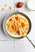 View from above of a bowl of cornflakes and strawberry slices in milk with a spoon resting in it. A fresh strawberry and a glass of milk stand next to it on the surface.