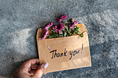 A hand holds an envelope with purple flowers and a handwritten thank you note against a textured grey background - a symbol of gratitude