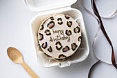 Top view of a mini bento birthday cake with a leopard print and a happy birthday message in a white box to take away, accompanied by a wooden spoon and a satin ribbon.