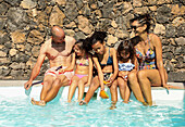 Eine lebhafte fünfköpfige Familie genießt einen sonnigen Tag am Pool. Sie sitzen mit den Füßen im Wasser, lächeln und haben Spaß in der strahlenden Sonne.