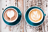 Two blue cups of cappuccino and hot chocolate with heart and leaf motifs stand on a weathered wooden table and create a cosy, rustic coffee house atmosphere.