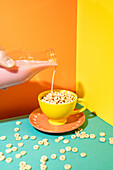 A dynamic and lively image of milk being poured into a bright yellow cup filled with cereal against a two-colour background of orange and yellow.