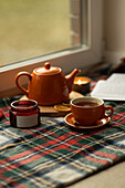 An inviting autumn scene shows an orange teapot and cup on a chequered blanket by the window, accompanied by a lit candle and dried orange slices, creating a warm and cosy atmosphere.