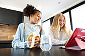 A smiling girl eating a banana and a young woman share a joyful moment in a bright, modern kitchen. They engage with a red table on the counter and enjoy a relaxed, informal activity.