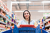 A young woman looks directly into the camera as she pushes a shopping trolley in a supermarket. She has a friendly smile and is wearing a casual pastel-coloured hoodie.