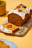 A freshly baked carrot cake with cream cheese icing, walnut pieces and aromatic rosemary, served on a rustic wooden board over a linen cloth