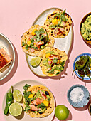 Blick von oben auf die farbenfrohen Tacos, die kreativ auf einem pastellrosa Hintergrund arrangiert sind. Zu den Zutaten gehören gegrillter Lachs, frischer Koriander, Mangoscheiben, Avocados und Limettenspalten, die eine verlockende und gesunde Mahlzeit darstellen.