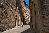 Sunlit cobblestone street in Volterra, Tuscany, Italy, lined with medieval stone buildings A woman strolls along the quiet alleyway, showing the town's rich history and warm atmosphere