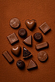 Top view of a variety of chocolate truffles artfully arranged on a background of rich cocoa powder. The collection includes heart-shaped, square and other beautifully moulded chocolates, perfect for dessert lovers.