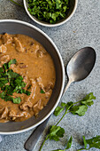 A tasty bowl of beef stroganoff with tender slices of meat in a creamy sauce, garnished with fresh coriander Another bowl contains freshly chopped coriander