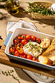 A dish of baked Camembert cheese, surrounded by cherry tomatoes and garlic, served with crusty bread. Garnished with herbs for a flavourful presentation.