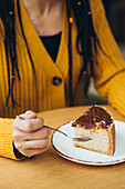 A person in a yellow jumper holds a fork and is ready to enjoy a delicious piece of cake on a white plate The cake is sprinkled with chocolate and nuts and stands on a wooden table