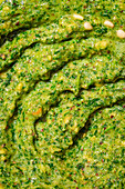 Nahaufnahme einer frisch zubereiteten Pesto-Sauce, deren leuchtend grüne Farbe und Textur mit Pinienkernen, Knoblauch und verschiedenen Kräutern gesprenkelt ist.