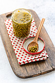 A jar of green, home-made organic peas stands on a rustic wooden board, accompanied by a wooden spoon on a dotted cloth. The scene creates a cosy, homely kitchen atmosphere.