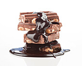 A stack of chocolate bars with almonds, drizzled with rich, shiny chocolate syrup on a white background. The luxurious texture and colour contrast create a tempting visual treat.