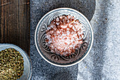 Close-up of pink Himalayan salt in a decorative ceramic bowl together with fresh green herbs on a rustic wood and stone background, perfect for healthy cooking inspiration.
