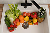 A variety of fresh fruit and vegetables, including tomatoes, oranges and pineapples, in a white kitchen sink, ready for washing. Healthy eating concept.