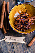 Top view of aniseed stars and cinnamon sticks in a textured yellow bowl on a wooden background, accompanied by a label with the inscription Spices