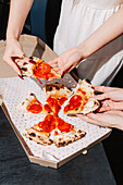 Friends share slices of wood-fired chilli and pepperoni pizza with salami, mozzarella and tomato sauce. Served in a takeaway box, perfect for a casual and fun meal together.