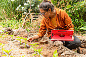 A woman with glasses and a red board carefully inspects the plants in a drip-irrigated garden surrounded by greenery and demonstrating sustainable cultivation methods