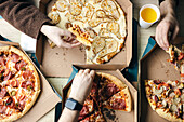View from above of a group of unrecognisable friends enjoying various pizzas together. Various hands, both male and female, reach into cardboard boxes to remove slices, showing a casual dining experience.