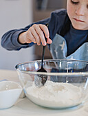 A child mixes flour in a glass bowl with a black spoon in a domestic kitchen. The scene shows a close-up of the activity, focussing on the child's hand and the baking ingredients.