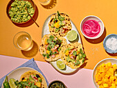 Draufsicht auf die Zubereitung einer Taco-Mahlzeit vor einem leuchtend gelben und rosa Hintergrund. Das Bild zeigt Tacos gefüllt mit Lachs, Mango, Koriander und Avocado. Als Beilagen sind Guacamole, eingelegte Zwiebeln, Limettenspalten und Salz zu sehen.
